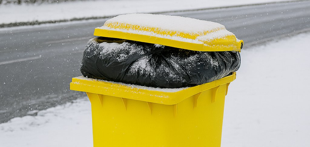 Gelbe Mülltonne am verschneiten Straßenrand, prall gefüllt mit schwarzen Müllsäcken. Der Deckel liegt schräg auf dem Müll auf.