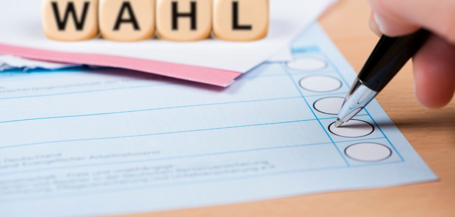 Hand hält Stift und markiert ein Feld auf einem Wahlzettel, daneben liegen farbige Unterlagen und oben stehen Würfel mit dem Wort WAHL.